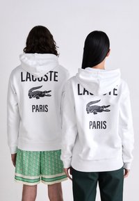 Du žmonės atsisuko nuo kameros, vilkintys baltas gaubtas su juodu Lacoste logotipu ir tekstu "PARIS"; vienas dėvi žalius raštuotus šortus, o kitas - tamsias kelnes.