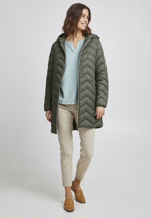 Cappotto imbottito acolor verde oliva con cappuccio, chiusura con zip, caratterizzato da cuciture a zigzag. Indossato sopra una maglietta azzurro chiaro, pantaloni beige e scarpe marroni.