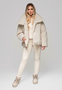 Béžová péřová bunda s oversized límcem, oblečená přes světlý top a úzké béžové legíny. Celkový vzhled doplňují béžové šněrovací kotníkové boty.