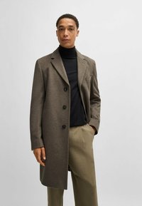 Cappotto in misto lana marrone con rever a punta, tre bottoni neri, taschino sul petto e maniche lunghe, indossato sopra un dolcevita nero.