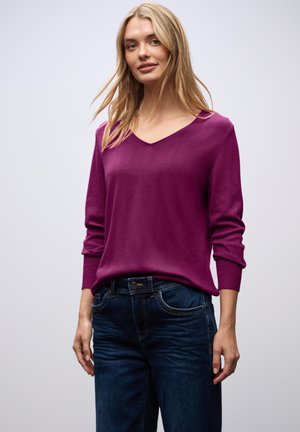 Enganliegender lila Strickpullover mit V-Ausschnitt und langen Ärmeln. Kombiniert mit dunkelblauen Jeans, die über normale Taschen und einen Metallknopf verfügen.
