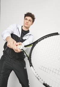 Černobílý sportovní outfit obsahující bundu s kapucí s geometrickými vzory a úzké kalhoty. Před sebou je držena tenisová raketa.