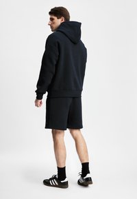 Mørkeblå hettegenser og shorts-sett laget av bomullsblanding. Har en avslappet passform, stor hette og sidelommer. Brukt sammen med svarte og hvite joggesko.