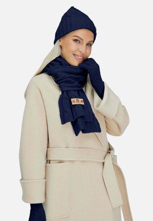 Marineblaue Strickmütze, Schal und Handschuhe-Set mit Zopfmuster, begleitet von einem cremefarbenen Mantel mit Gürtel und seitlichen Taschen.