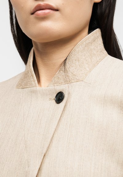 Close-up af en person iført en beige frakke med høj krave og en enkelt synlig sort knap nær halsen.