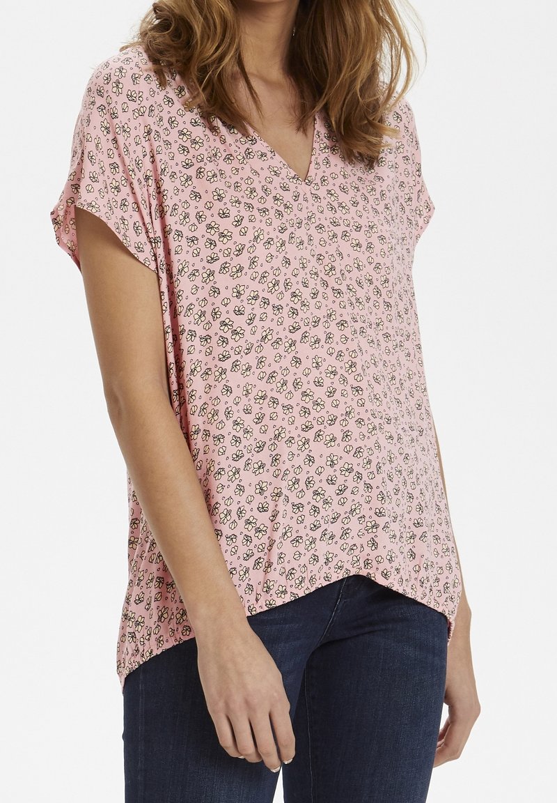 Le haut rose à manches courtes présente un col en V avec un motif floral noir et blanc, fabriqué en tissu léger avec des fentes sur les côtés.