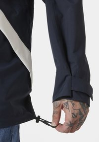 Marineblauwe jas met een witte diagonale streep, voorzien van een verstelbare trekkoorden aan de onderrand en een textuurstof. Hand met tatoeages die het trekkoord vasthoudt.