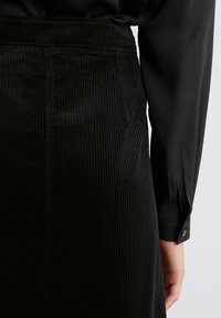 Jupe en velours côtelé noir avec des côtes verticales, stylisée avec une chemise noire à manches longues. Elle présente une taille et des poignets à boutons.