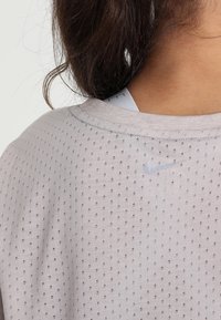 Camiseta atlética de color gris claro que absorbe la humedad, con pequeñas perforaciones de malla y un logo de Nike blanco cerca del cuello. Textura suave y corte relajado.