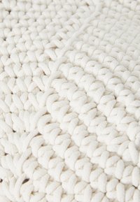 Tappeto di cotone lavorato a maglia di colore bianco sporco con filato spesso e testurizzato, caratterizzato da un motivo geometrico creato da trecce e anelli intrecciati.