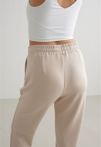 Beige Jogginghosen mit elastischem Bund und Seitentaschen, aus einem weichen Stoff, kombiniert mit einem weißen, taillierten Tanktop. Glatte Textur, minimalistisches Design.