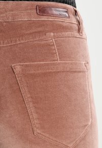 Světle růžové kordové kalhoty s výraznou zadní kapsou a koženou značkovou etiketou. Textura je jemná a hladká, s decentními švy.