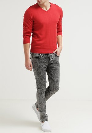 Rotes V-Ausschnitt-Strickoberteil kombiniert mit grauen Acid-Wash-Skinny-Jeans und weißen Slip-On-Schuhen. Der Stoff wirkt glatt, mit figurbetonten Designdetails.