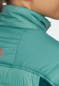 Turkusowa kurtka ocieplana z wysokim kołnierzem, o gładkiej fakturze z kontrastowym czerwonym logo na ramieniu.