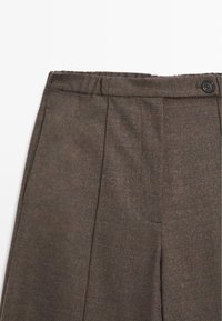 Gros plan de la ceinture d’un pantalon en laine marron avec fermeture par bouton et poches latérales, montrant la texture du tissu et les détails des coutures.