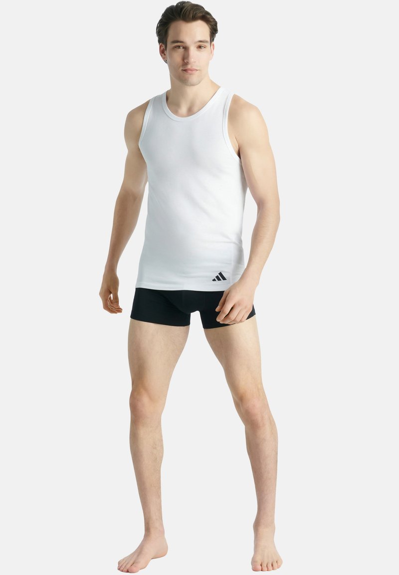 Bílý bavlněný top bez rukávů s širokými ramínky, spárovaný s černým spodním prádlem. Obsahuje černé logo Adidas v dolním levém rohu topu.