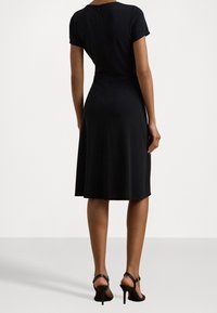 Zwarte jurk met korte mouwen en een getailleerde taille, die uitloopt in een knielange rok. Gladde textuur met een rits aan de achterkant.