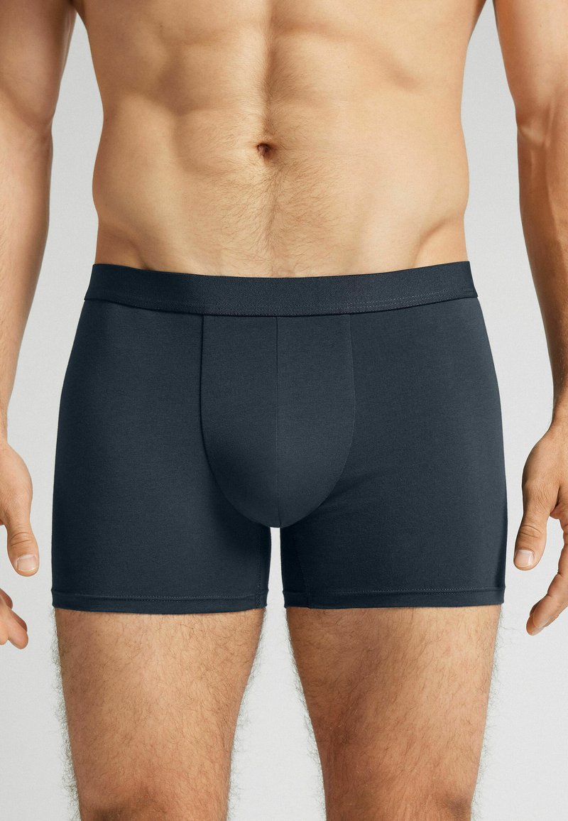 Mörkgrå herrboxershorts, tillverkade av mjukt tyg, med en åtsittande design, elastisk midjeband och en kort längd som når till mitten av låret.