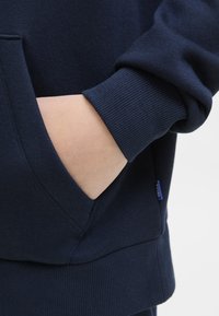 Mano parcialmente dentro del bolsillo frontal de una sudadera azul marino con puños y dobladillo acanalados.