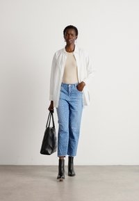 Hvit oversized skjorte, beige topp, blå jeans med svake mønstre, svarte ankelsko, og en svart vevd håndbag. Enkel bakgrunn.