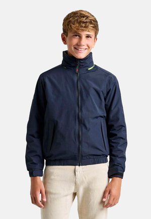 Chaqueta azul marino con cuello alto, cierre de cremallera y dobladillo elástico. Presenta acentos en verde y dos bolsillos frontales. Textura suave.