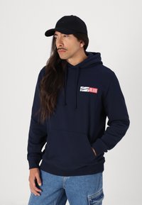 Marineblå hettegenser med frontlomme, logopatch i rødt og hvitt. Kombinert med lyseblå cargo-jeans, båret med en svart caps.