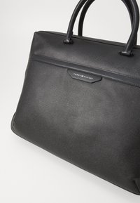 Sac à main en cuir noir texturé avec deux poignées supérieures et une forme rectangulaire. Présente un logo embossé subtil sur une accent en cuir noir.