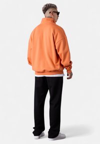Oranger Fleece-Pullover mit hohem Kragen, lockerer Passform und gerippten Bündchen, kombiniert mit schwarzen Sweatpants und weißen Sneakers.