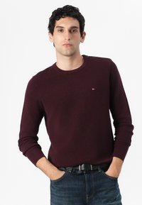 Pull en maille bordeaux avec un col rond et des manches longues, arborant un petit logo sur la poitrine, associé à un jean en denim foncé.