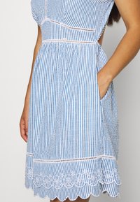 Sininen ja valkoinen raidallinen mekko, jossa on vartalonmyötäinen yläosa, volyymikas hame, taskut ja aallotettu pitsireunus helmassa. Valmistettu kevyestä kankaasta.
