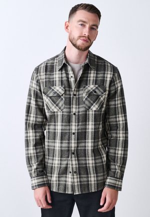 Ung mand iført en sort-hvid ternet skjorte med knapper og klaplommer på brystet, står foran en ensfarvet lys baggrund.