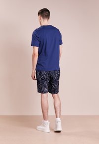 Granatowa bawełniana koszulka zestawiona z szortami w ciemnoniebieski, skropionym wzorem, z białymi akcentami. Model nosi białe sneakersy za kostkę.