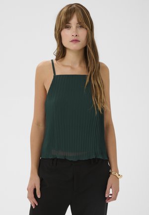 Femme portant un haut sans manches plissé vert foncé à fines bretelles et un pantalon noir, debout devant un fond clair uni.