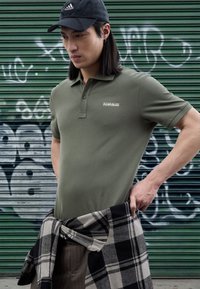 Oljčno zelena polo majica o tesnem kroju z majhnim logotipom, kombinirana s črno-sivim karo flanelom, privezanim okoli pasu, na ozadju grafita.