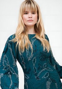 Teal kjole med blomsterperlebroderi og paljetter, med lange puffærmer og rund halsudskæring. Glat stofstruktur.