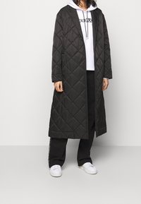 Lungo cappotto nero trapuntato con un motivo a rombi, davanti aperto e due tasche laterali, indossato sopra un hoodie bianco e pantaloni neri.