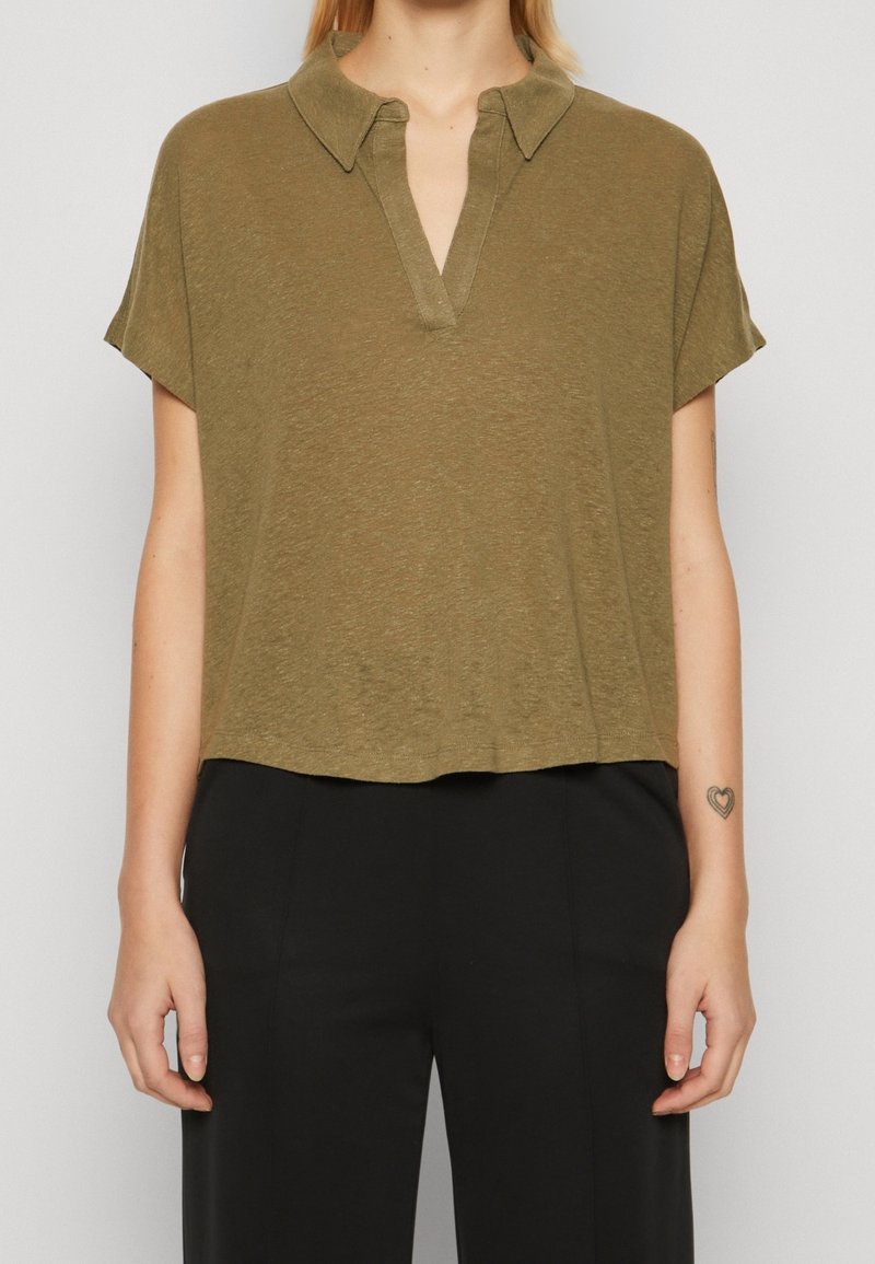 Femme portant un polo ample à manches courtes de couleur vert olive avec un pantalon noir, affichant un petit tatouage en forme de cœur sur son avant-bras gauche.