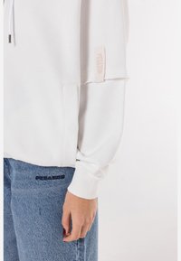 Felpa bianca con una vestibilità rilassata e polsini a coste, con un'etichetta sulla manica. Abbinata a jeans in denim blu chiaro, con il nome del marchio ricamato.