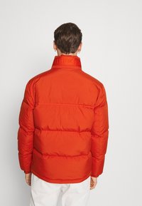 Man med kort brunt hår som bär en ljusorange pufferjacka och vita byxor, sedd bakifrån mot en enkel bakgrund.