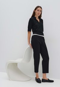 Schwarzes, kurzärmeliges Oberteil mit Kragen, kombiniert mit schwarzen, taillierten Hosen. Das Model steht neben einem gebogenen weißen Stuhl. Eleganter, minimalistischer Look.