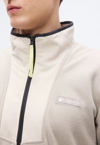 Μπεζ φλις μπουφάν με μαύρο φερμουάρ και γιακά, με μικρό λογότυπο Columbia στο αριστερό στήθος. Η τσόχινη λαβή του φερμουάρ έχει έντονη κίτρινη απόχρωση.