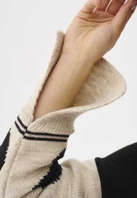 Pull en tricot beige et noir avec un col prominent rabattu, présentant des rayures noires et une finition texturée. Main partiellement visible.