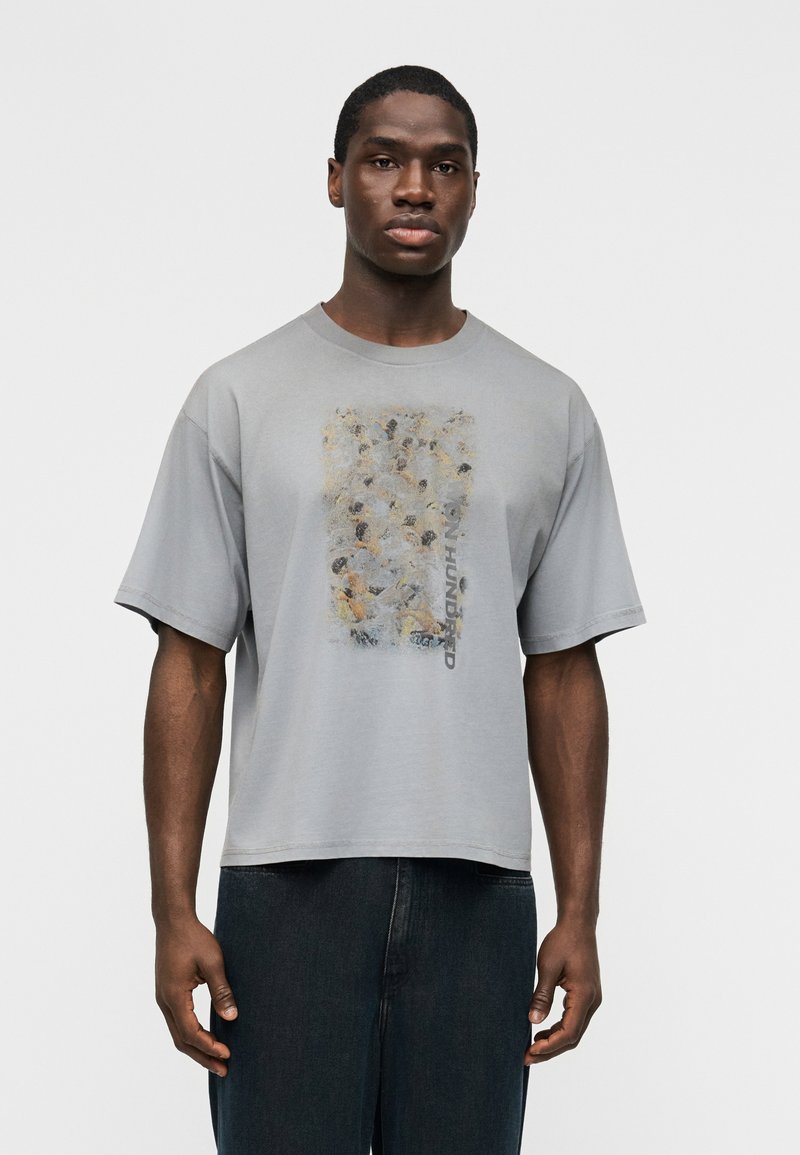 Άνδρας που φοράει φαρδιά γκρι μπλο ύζα με αφηρημένο σχέδιο και κατακόρυφο κείμενο "Only Hundred", σε συνδυασμό με σκούρο τζιν, στέκεται μπροστά σε λευκό φόντο.