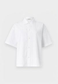 Λευκό πουκάμισο με κοντά μανίκια, κουμπιά και γιακά, με κάθετες πτυχώσεις μπροστά, παρουσιασμένο σε απλό φωτεινό φόντο.