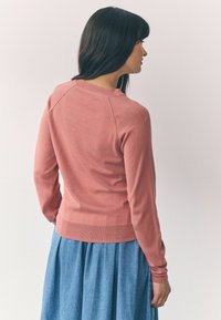 Maglione rosa chiaro con maniche lunghe, polsini a coste e colletto rotondo; indossato sopra una gonna plissettata blu; texture liscia.
