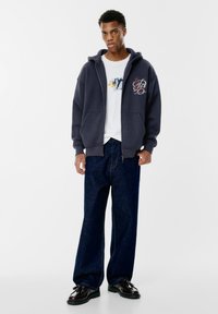 Tânărul stă în picioare, purtând un hanorac cu fermoar de culoare albastru închis, un tricou alb cu grafică, blugi largi din denim închis la culoare și mocasini negri cu ciucuri, pe un fundal alb.