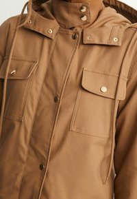 Brun lærredjakke med hætte, prydet med guldhardware, to frontlommer og knaplukning. Struktureret materiale med en glat overflade.
