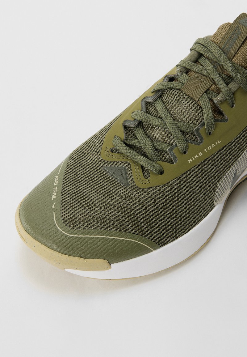Zapatilla deportiva verde oliva con parte superior de malla, cordones texturizados y suela de color marrón claro. Presenta una marca en el lateral y puntera reforzada.