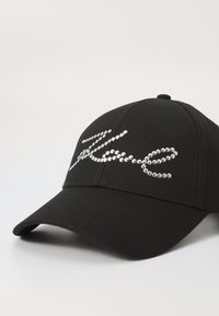 Melna beisbola cepure, izgatavota no auduma, ar spīdīgu rhinestone dizainu slīpā burtveidā uz priekšpuses. Ieliekta maliņa, strukturēta forma.