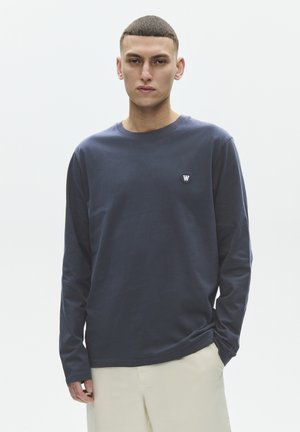 Ung mand med kort hår iført en mørkeblå langærmet skjorte med en lille logo på brystet og lyse bukser, stående neutralt.